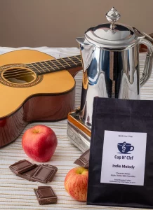 Indie Melody coffee bag placed next to a guitar, apples, and chocolate, evoking a sense of warmth and sweet flavor.