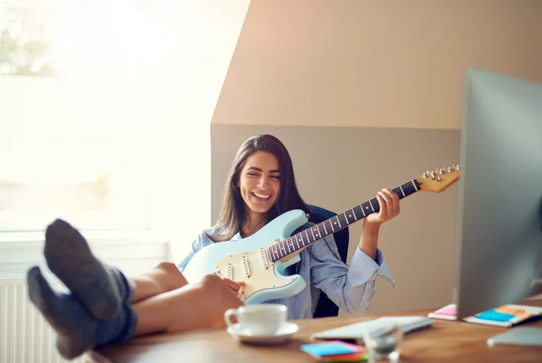 Woman playing an electric guitar while relaxing at her desk with a coffee cup.